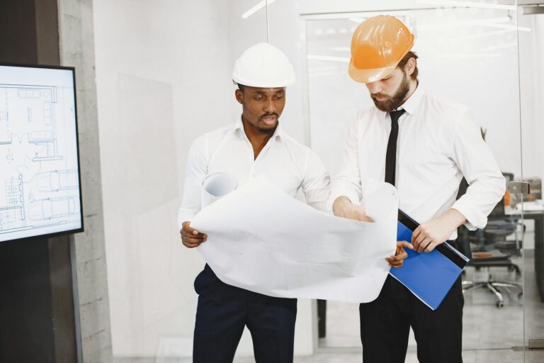 Two architects in hard hats review blueprints during a collaborative meeting in an office setting.