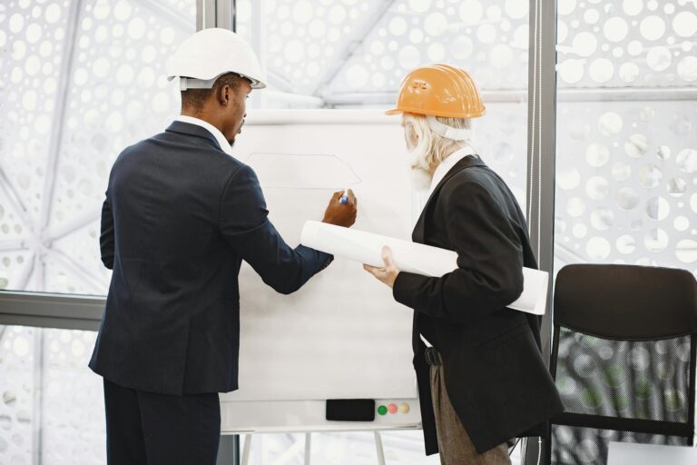 Two architects in hard hats working on a blueprint indoors, highlighting teamwork.